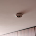 White circular smoke detector mounted on a pale pink ceiling in a hallway or room.