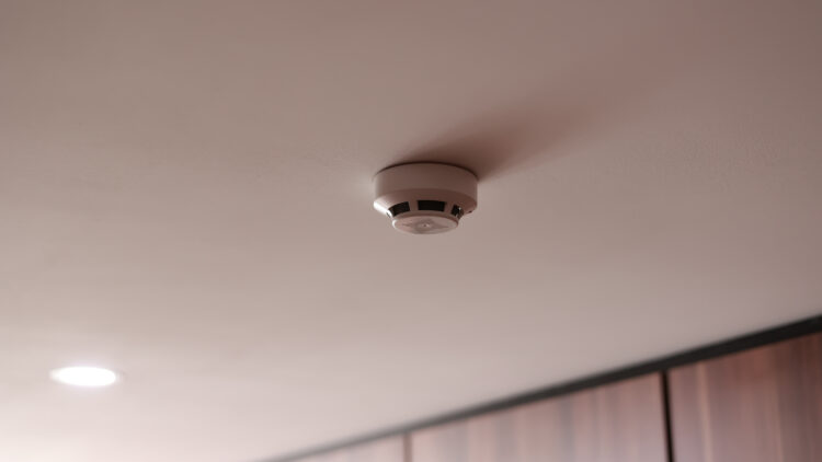 White circular smoke detector mounted on a pale pink ceiling in a hallway or room.