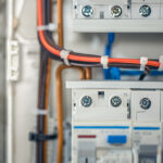 Close-up of an electrical breaker panel with red and black wires tied by white zip ties and labeled terminals (N) and numbered positions.