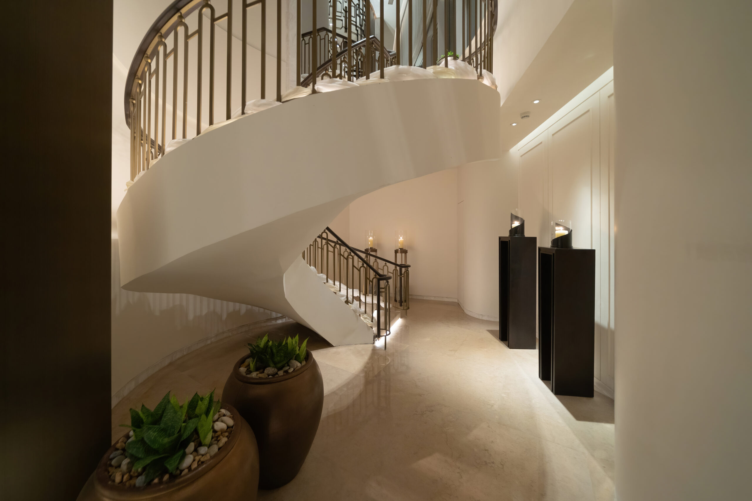 Elegant curved white spiral staircase inside a modern lobby, with metal railings and warm candlelight along the wall.