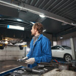 Auto mechanic in blue coveralls inspecting a car lifted on a hydraulic lift, holding a tool in a bright workshop.