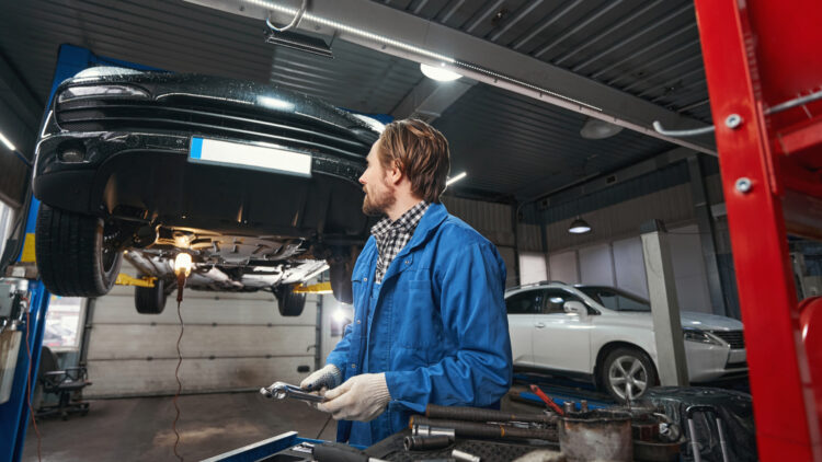 Auto mechanic in blue coveralls inspecting a car lifted on a hydraulic lift, holding a tool in a bright workshop.