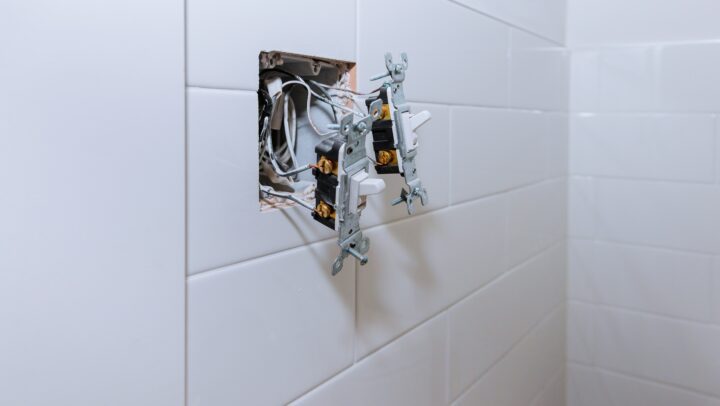 Exposed electrical wiring and two switches protruding from an open wall box in a tiled bathroom.