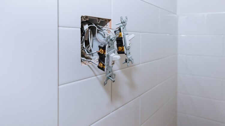 Exposed electrical wiring and two switches protruding from an open wall box in a tiled bathroom.